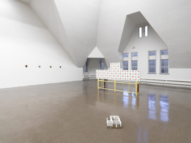 A stand of yellow cherrywood mounted on a wall with a small white, red and aluminum sculpture on top of it