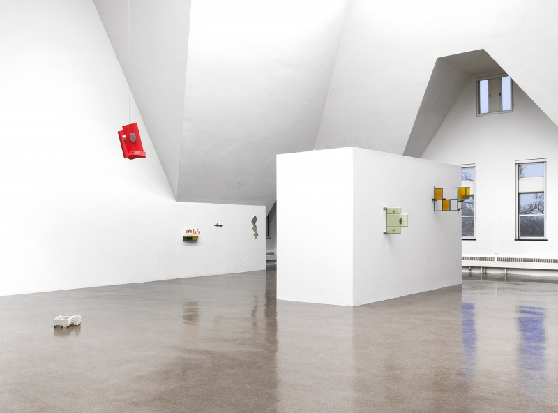 A stand of yellow cherrywood mounted on a wall with a small white, red and aluminum sculpture on top of it