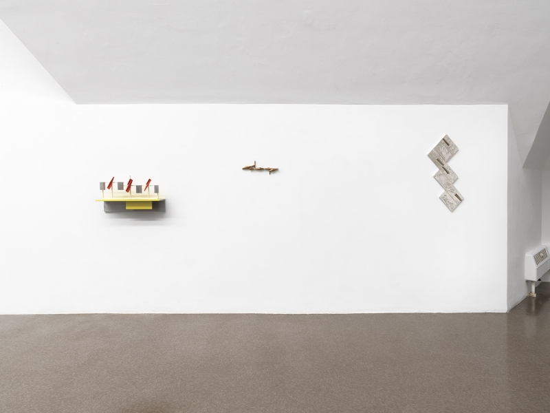 A stand of yellow cherrywood mounted on a wall with a small white, red and aluminum sculpture on top of it