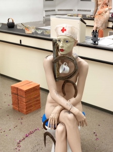 A mannequin sits on a toilet seat, wearing a nurse's hat, with a coffee cup and horseshoes dangling from her neck