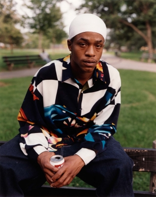 A Black man with a white head covering sits on a park bench holding a drink can.