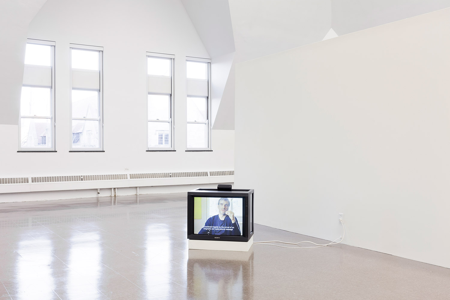 A TV resting on the floor plays a film of a man sitting in front of a window and talking.