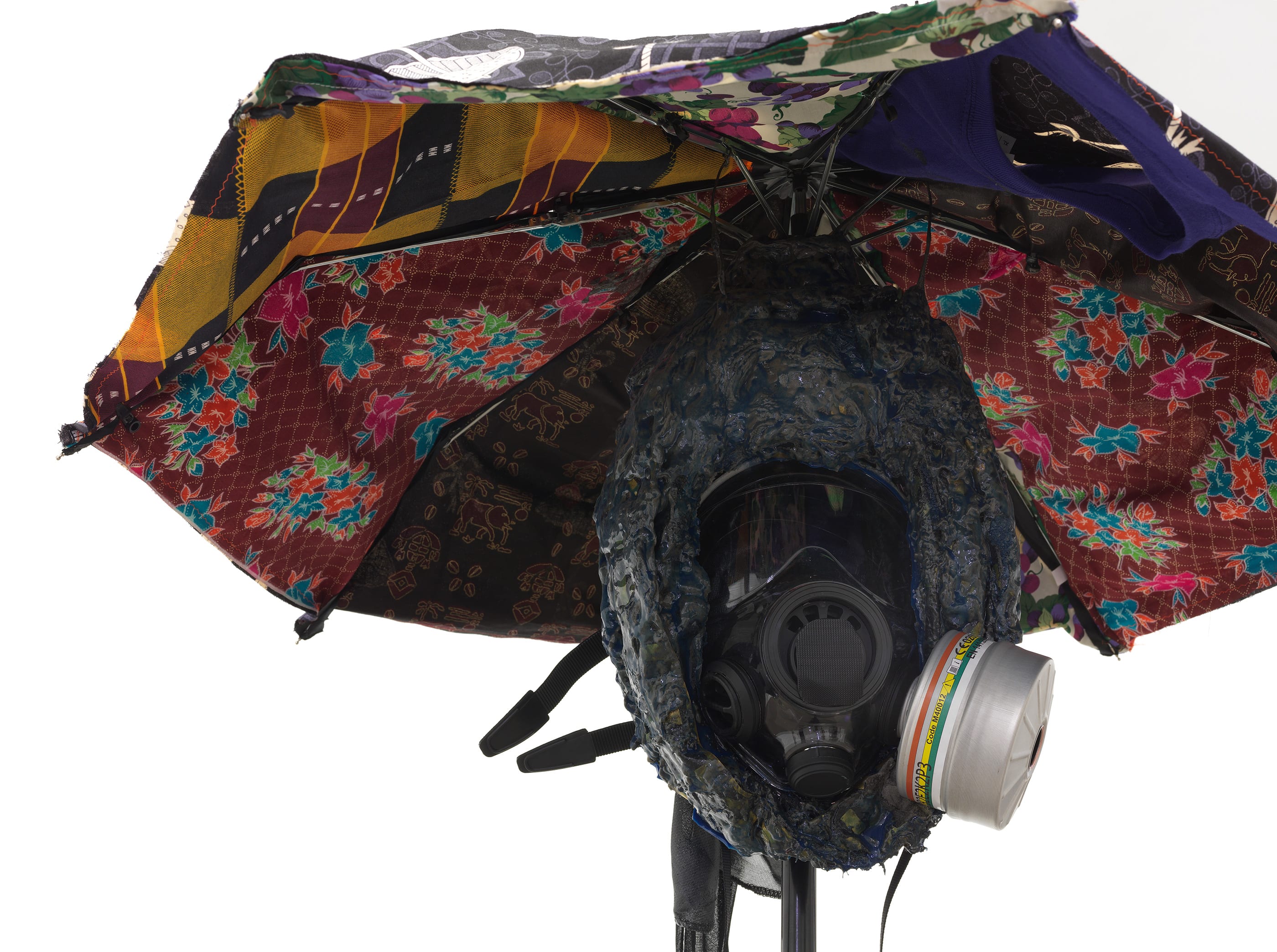 A patchwork umbrella covers a dark, head-shaped sculpture, which itself covers a gas mask.
